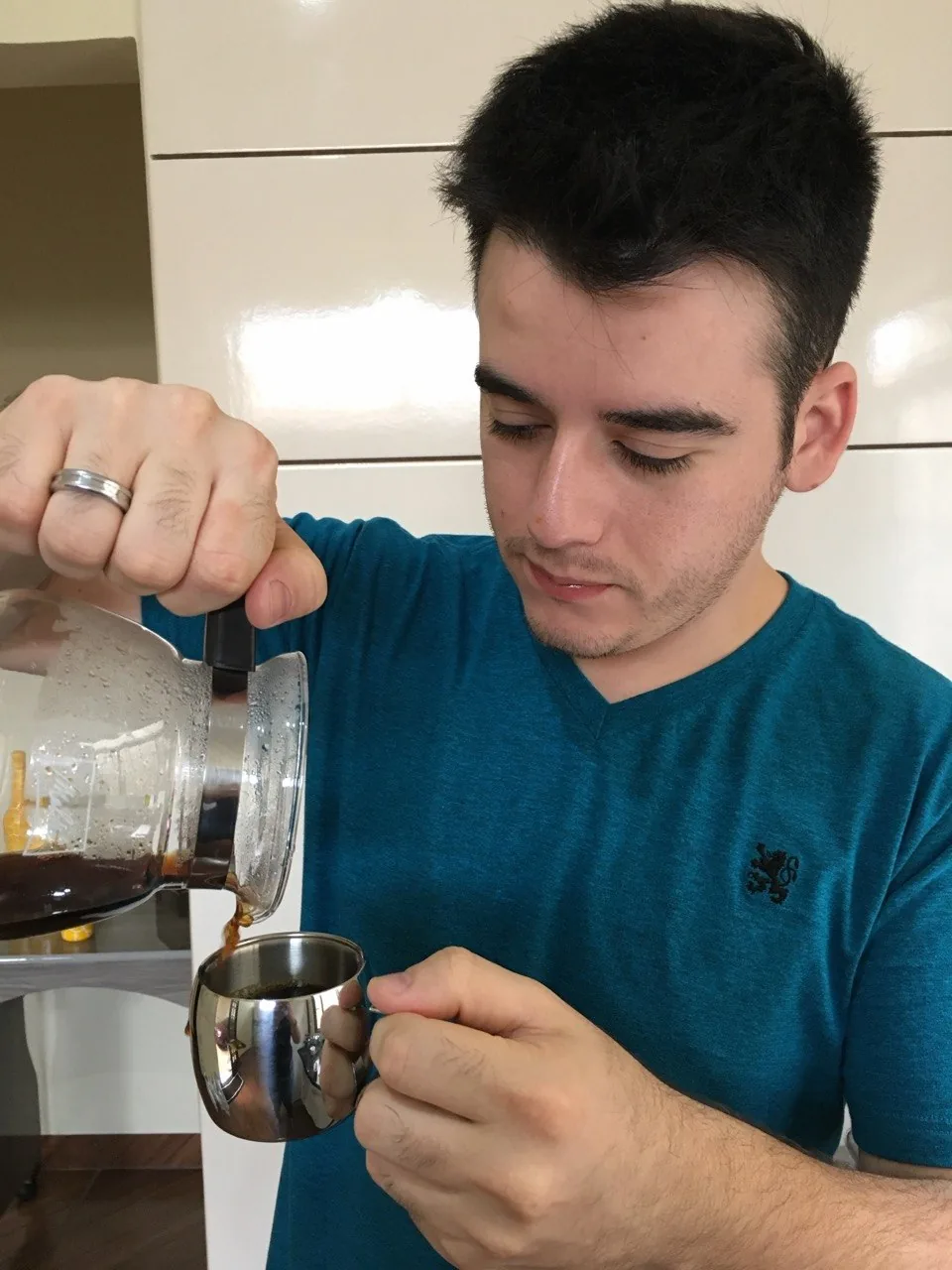Pessoa de camiseta azul servindo café de uma jarra de vidro em uma xícara de metal.