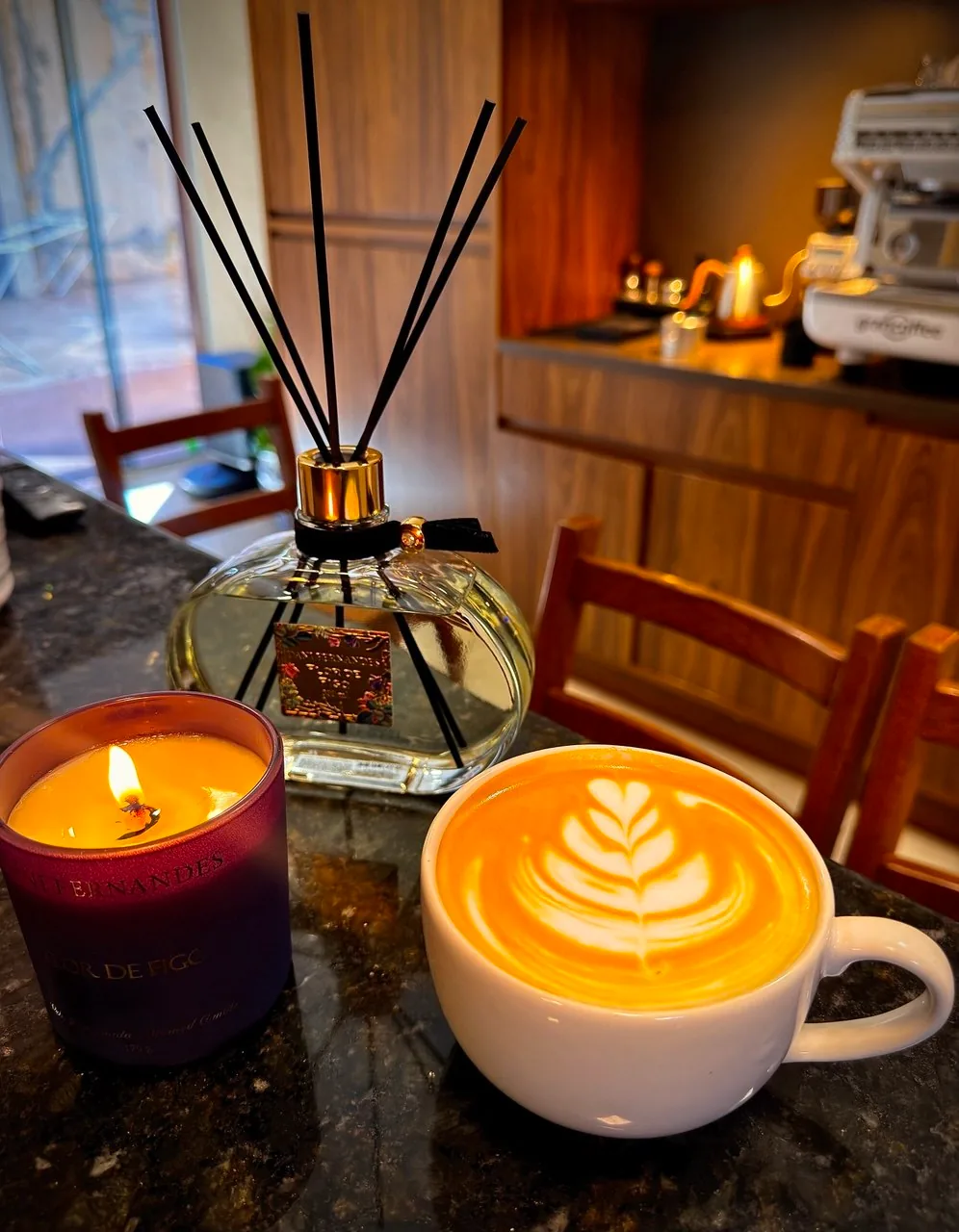 Xícara com latte art ao lado de uma vela acesa e um difusor, com a bancada de café ao fundo.