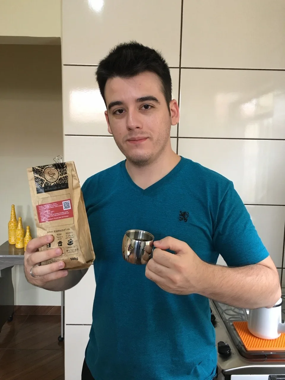 Pessoa de camiseta azul segurando um pacote de café Grão Gourmet e uma xícara de metal.