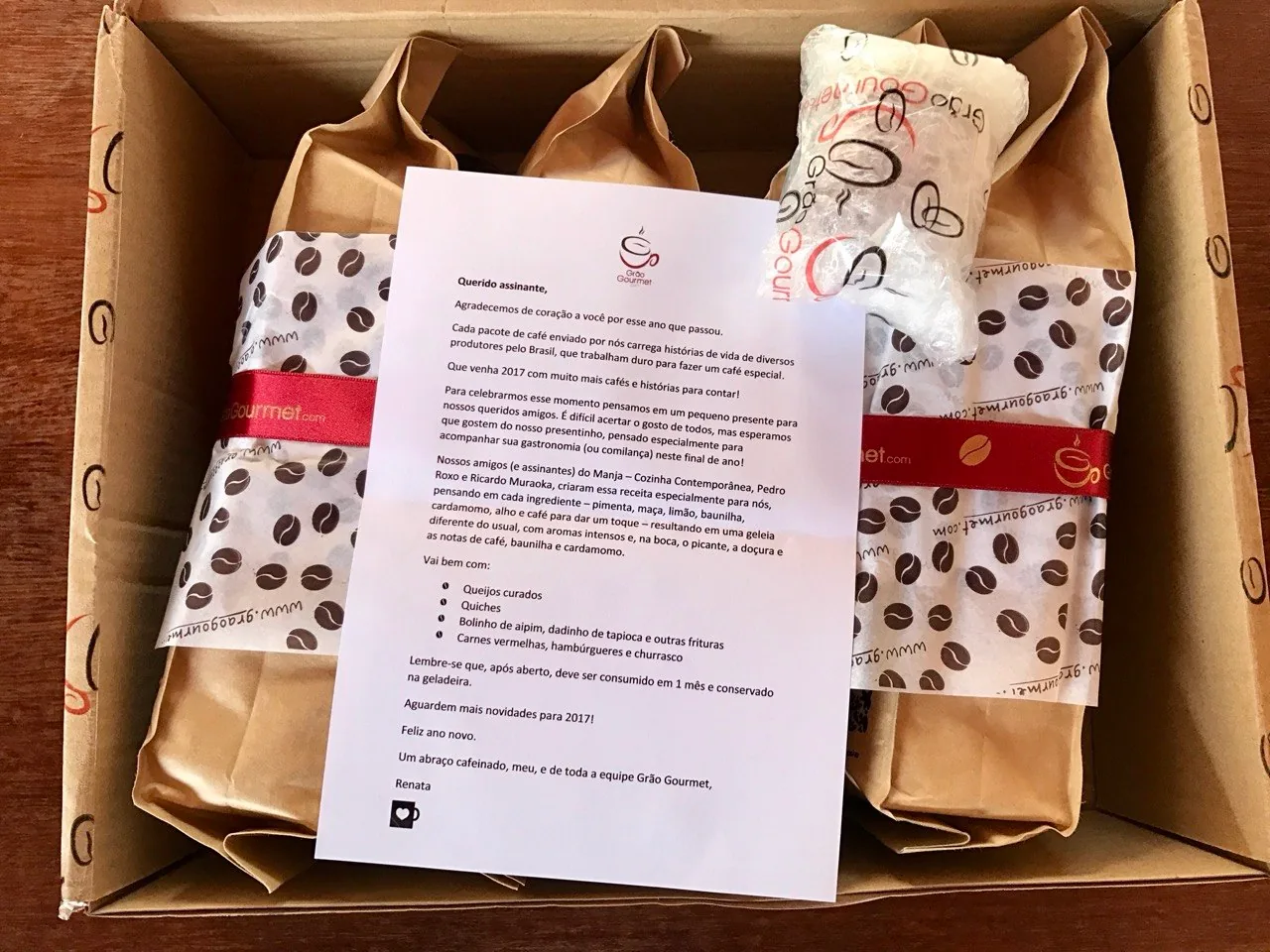 Caixa da Grão Gourmet com pacotes de café, um presente embalado e uma carta impressa.