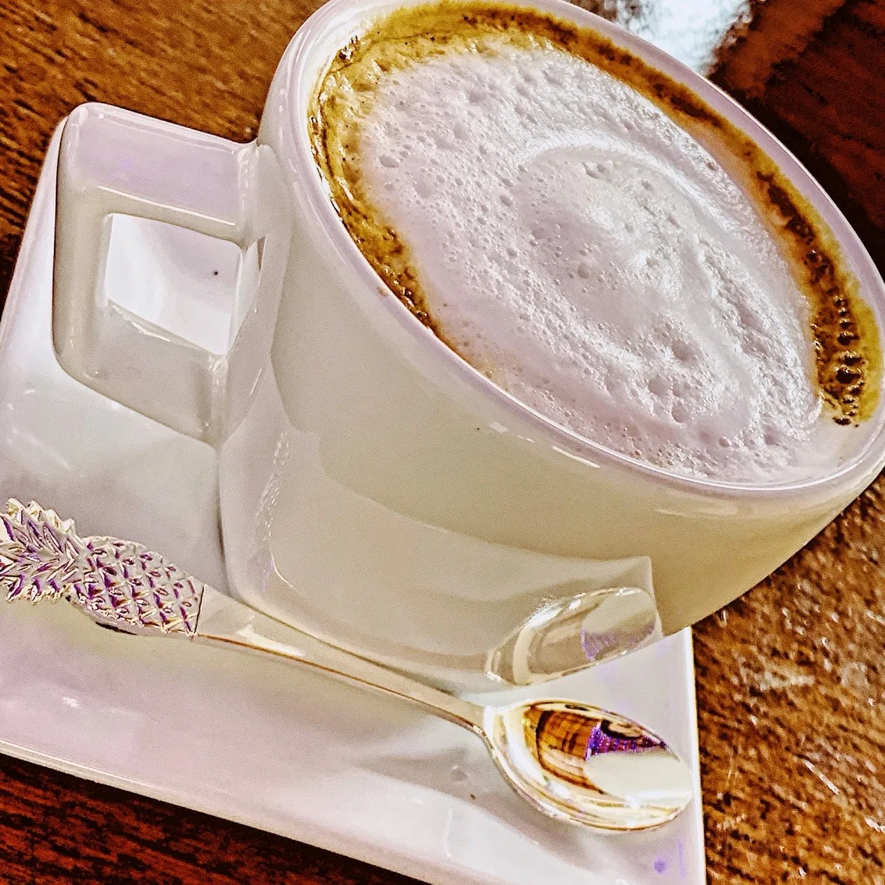 Xícara branca com café e uma camada espessa de espuma de leite, sobre pires com colher.