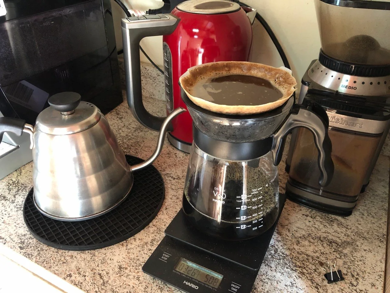 Jarra Hario V60 com coador sobre a balança, ao lado de uma chaleira de bico fino e um moinho.