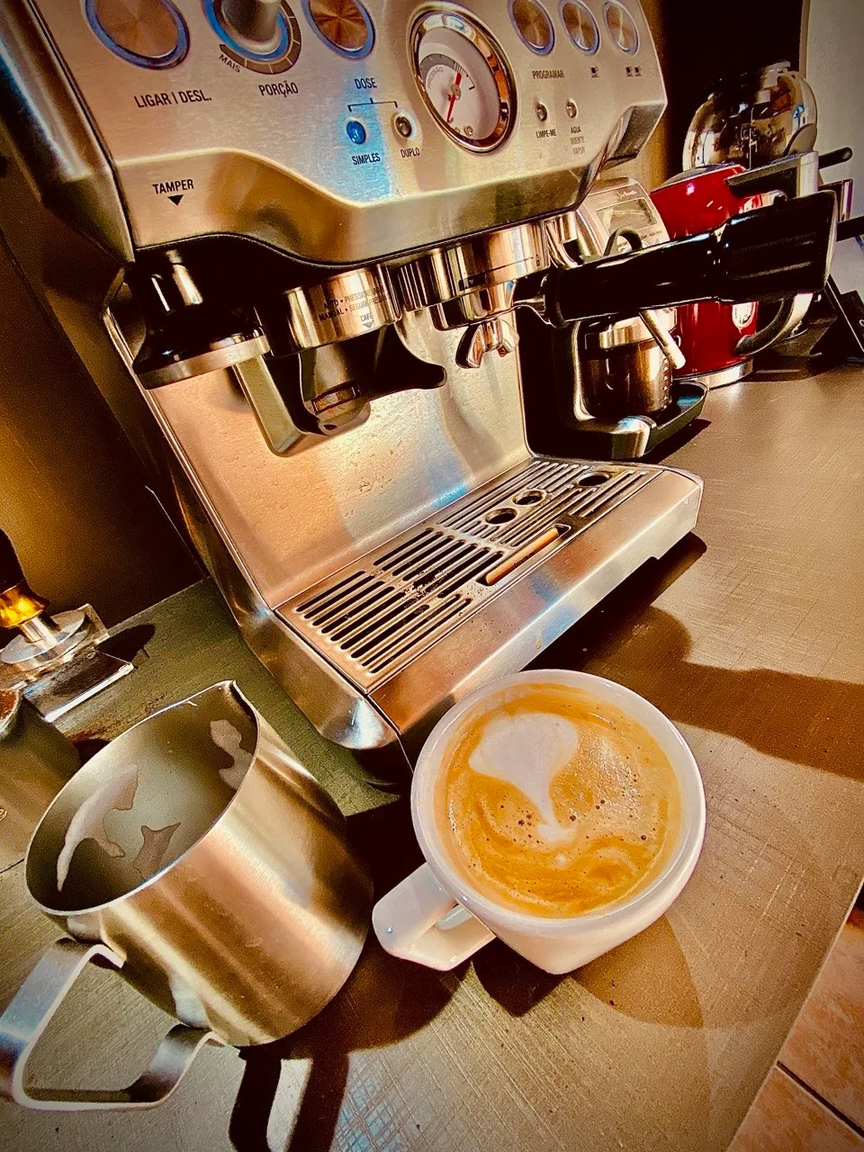 Máquina de espresso de inox com moinho integrado, jarra de leite e xícara de café com leite.
