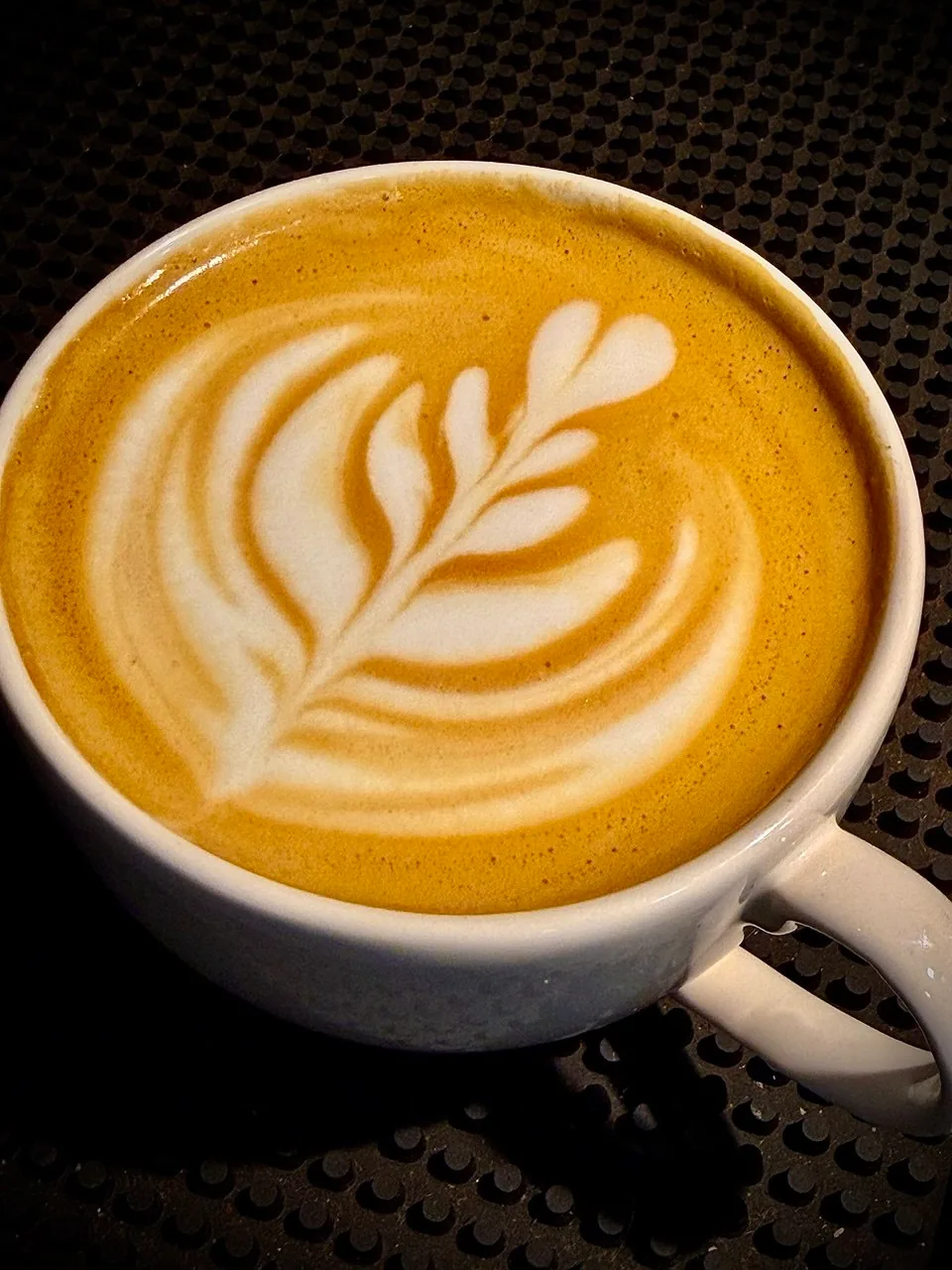 Xícara de café com latte art em formato de ramo apoiada sobre um tapete preto.