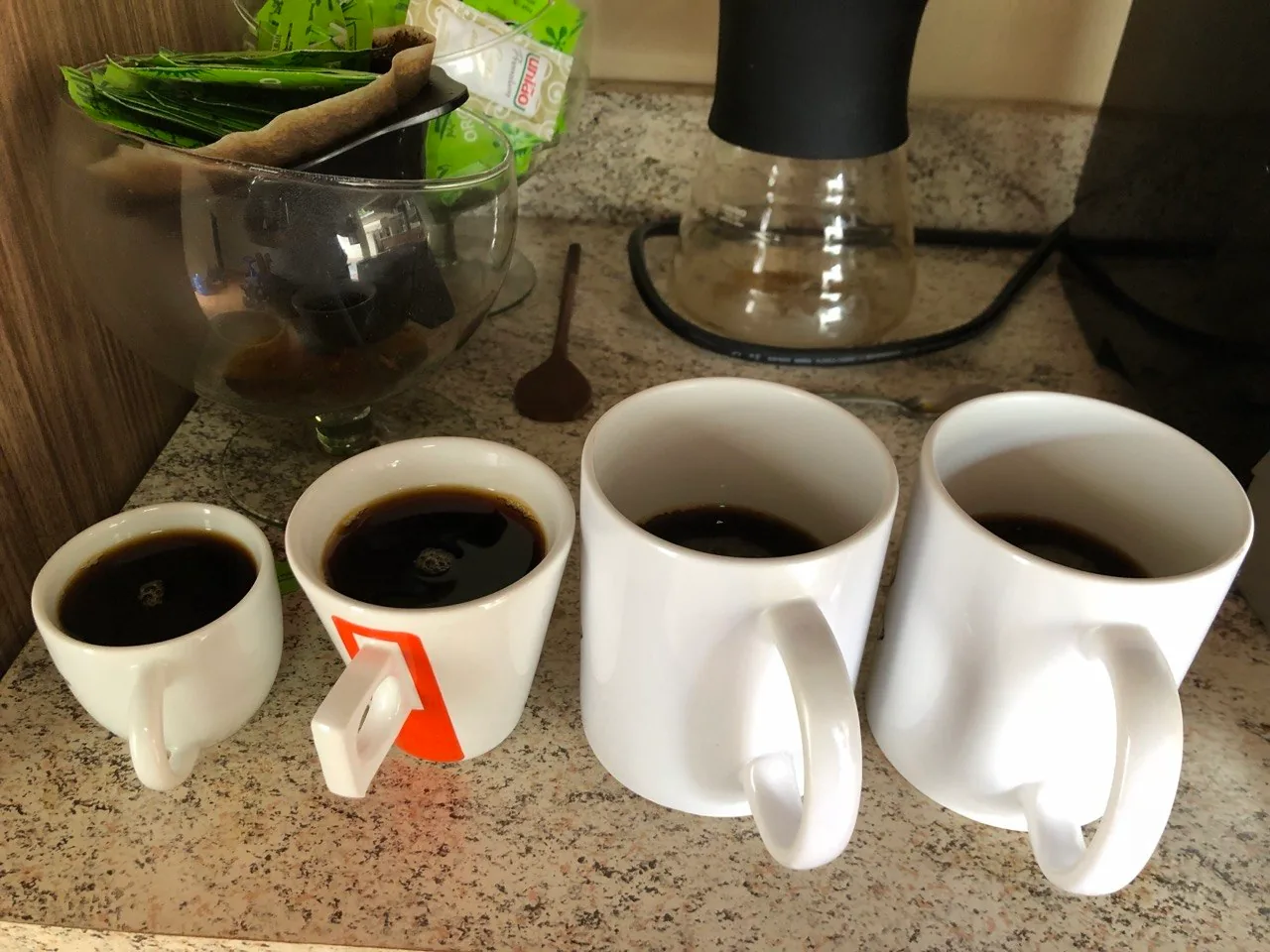 Quatro xícaras e canecas brancas de tamanhos diferentes, com café, alinhadas na bancada.