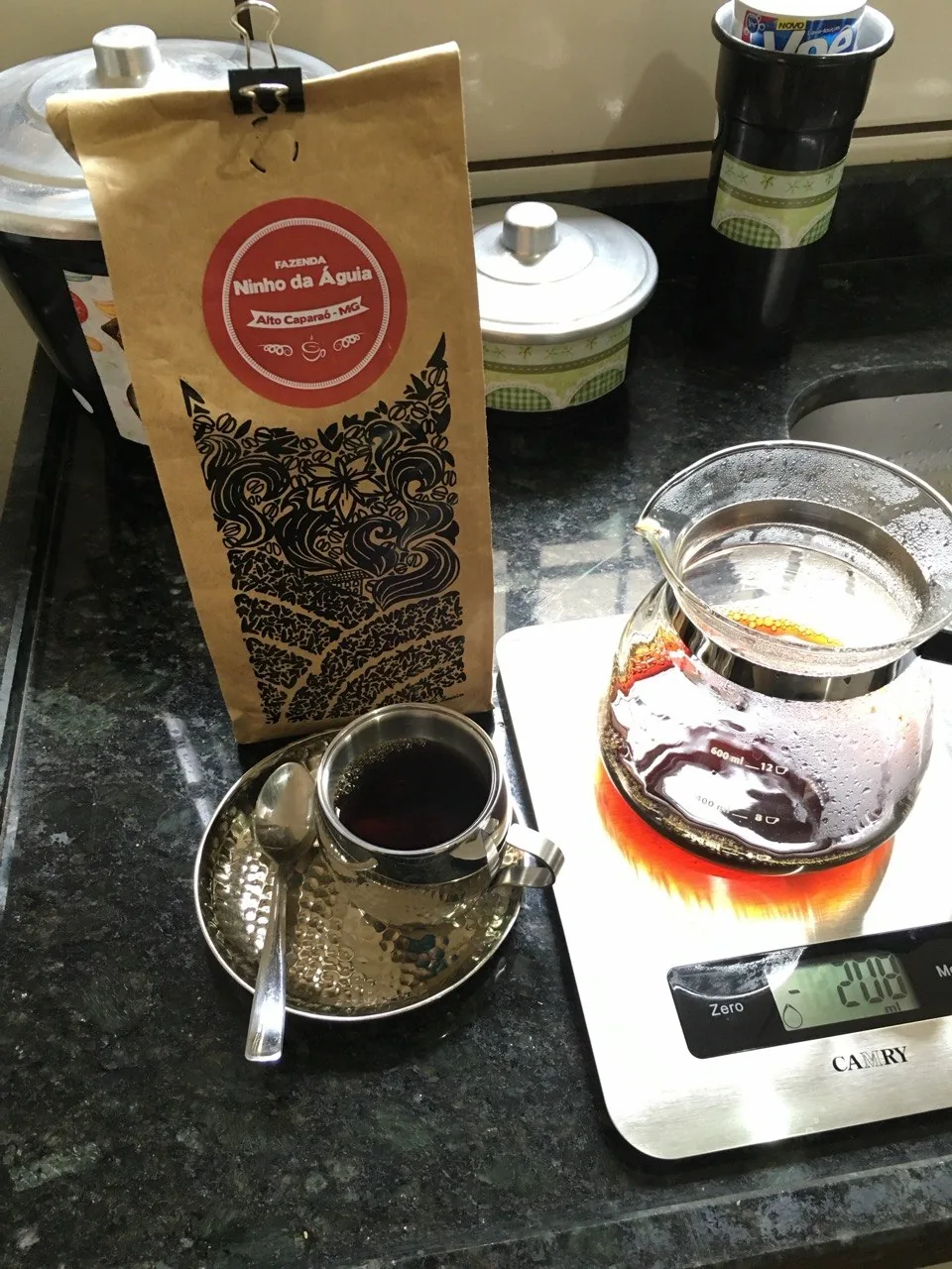 Pacote de café Ninho da Águia ao lado de uma xícara de metal e uma jarra com café sobre a balança.
