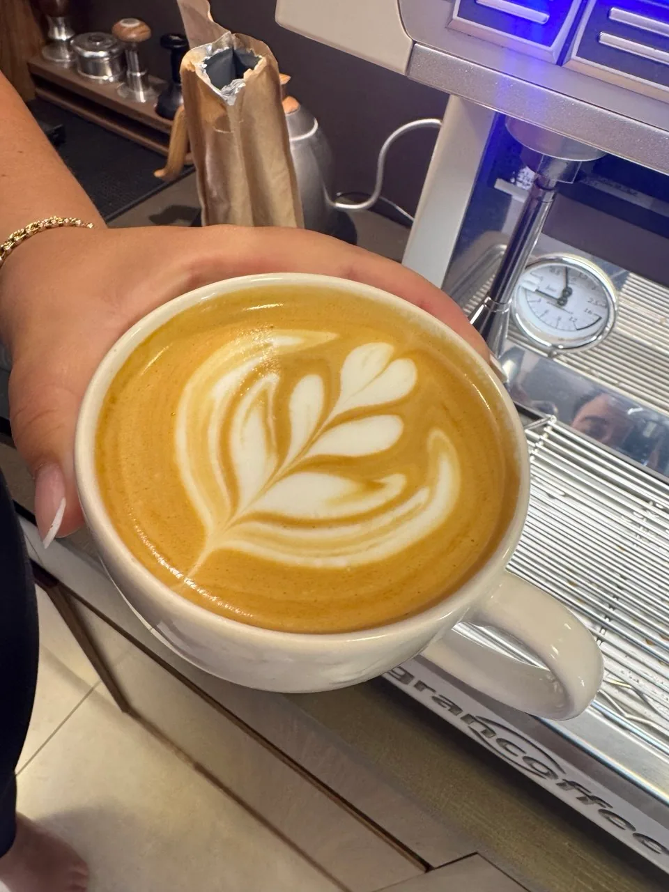 Mão segurando uma xícara de latte art diante de uma máquina de espresso de inox.