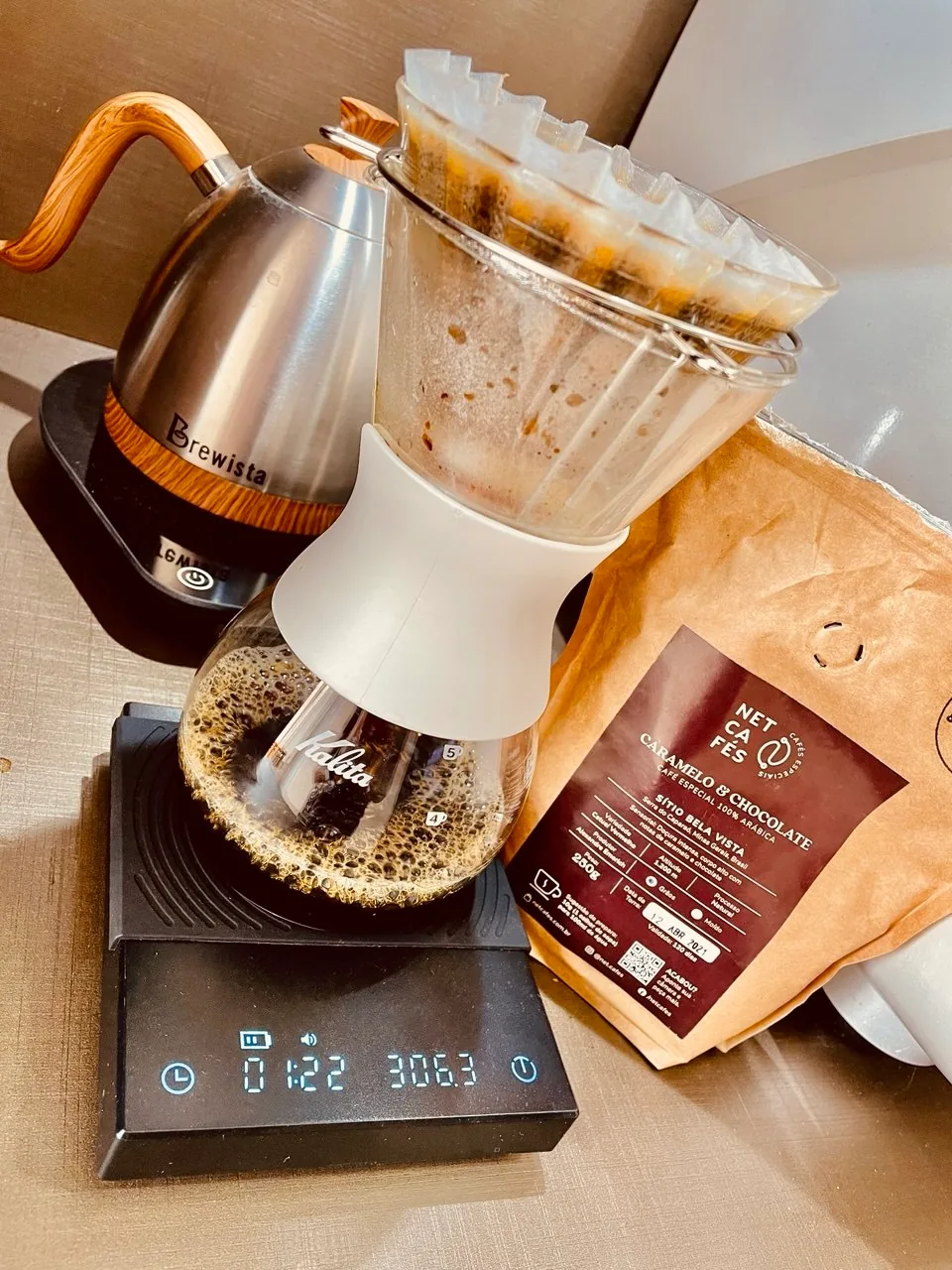 Coador de vidro sobre jarra Kalita e balança, com chaleira Brewista e pacote da Netcafés ao lado.