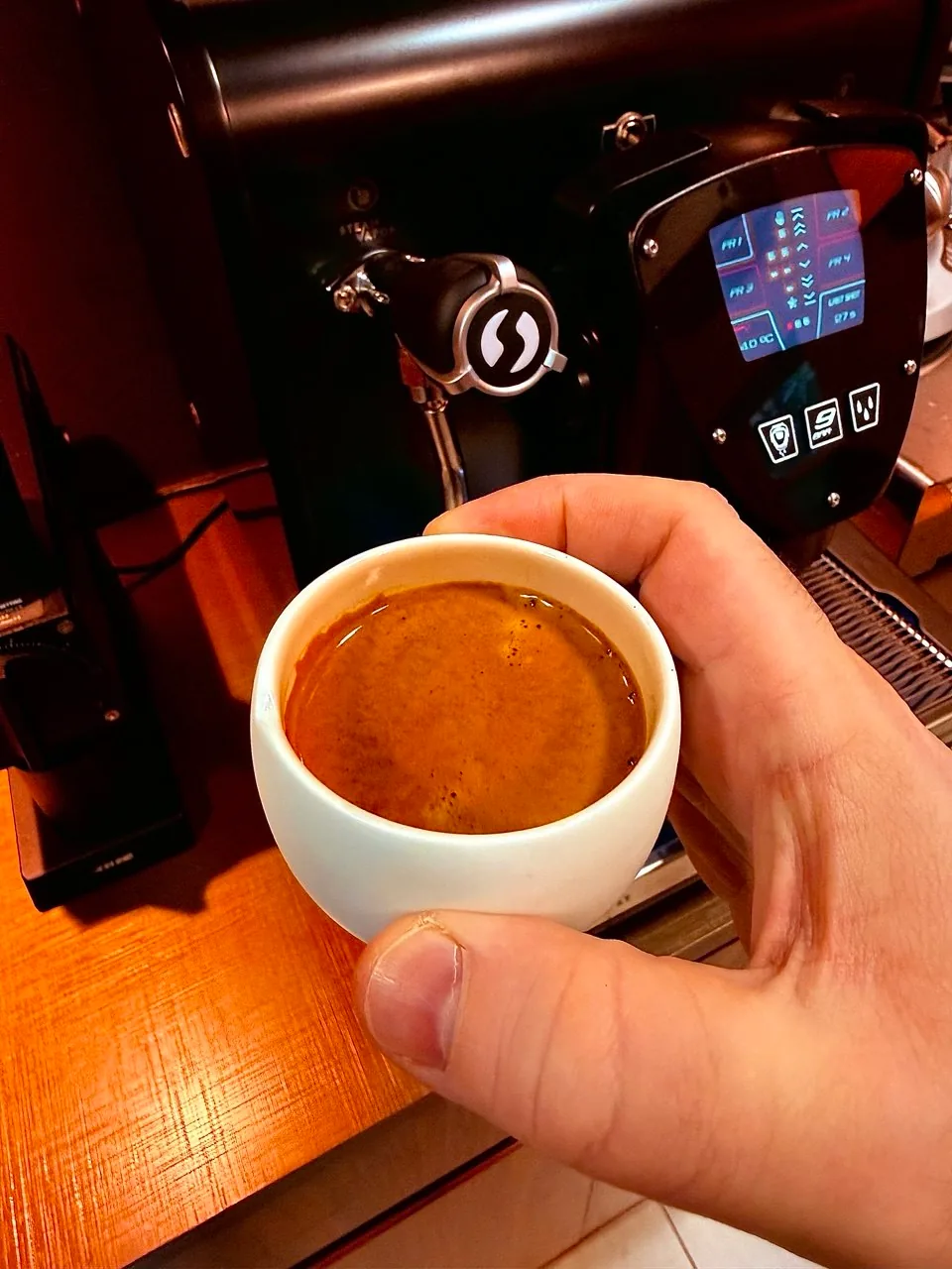 Mão segurando uma pequena xícara branca de espresso diante da máquina Fiamma.