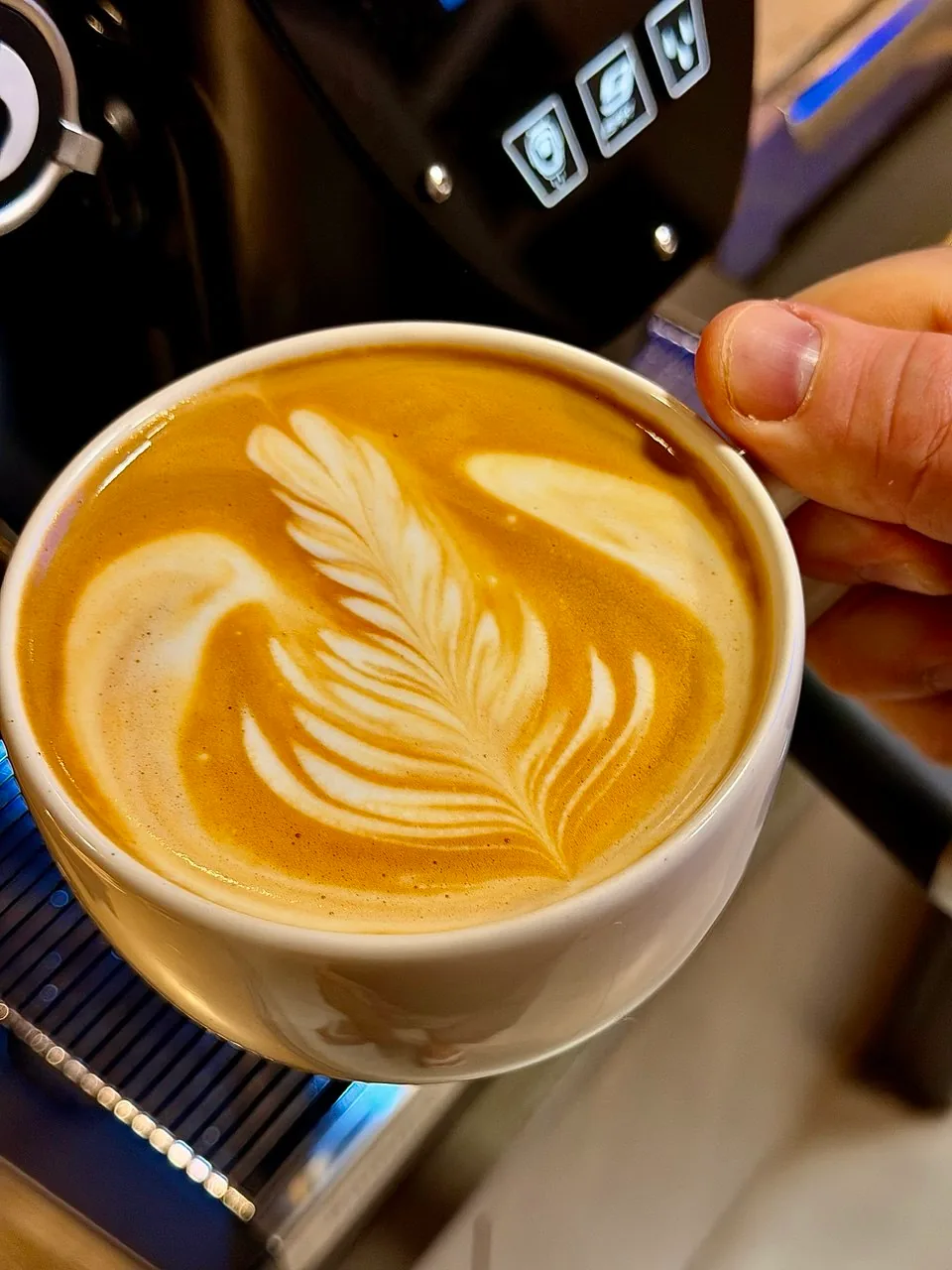 Xícara branca com latte art em formato de folha, segurada diante da máquina de espresso.