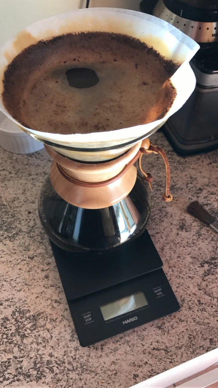 Cafeteira de vidro com colar de madeira e filtro de papel, apoiada sobre uma balança Hario.