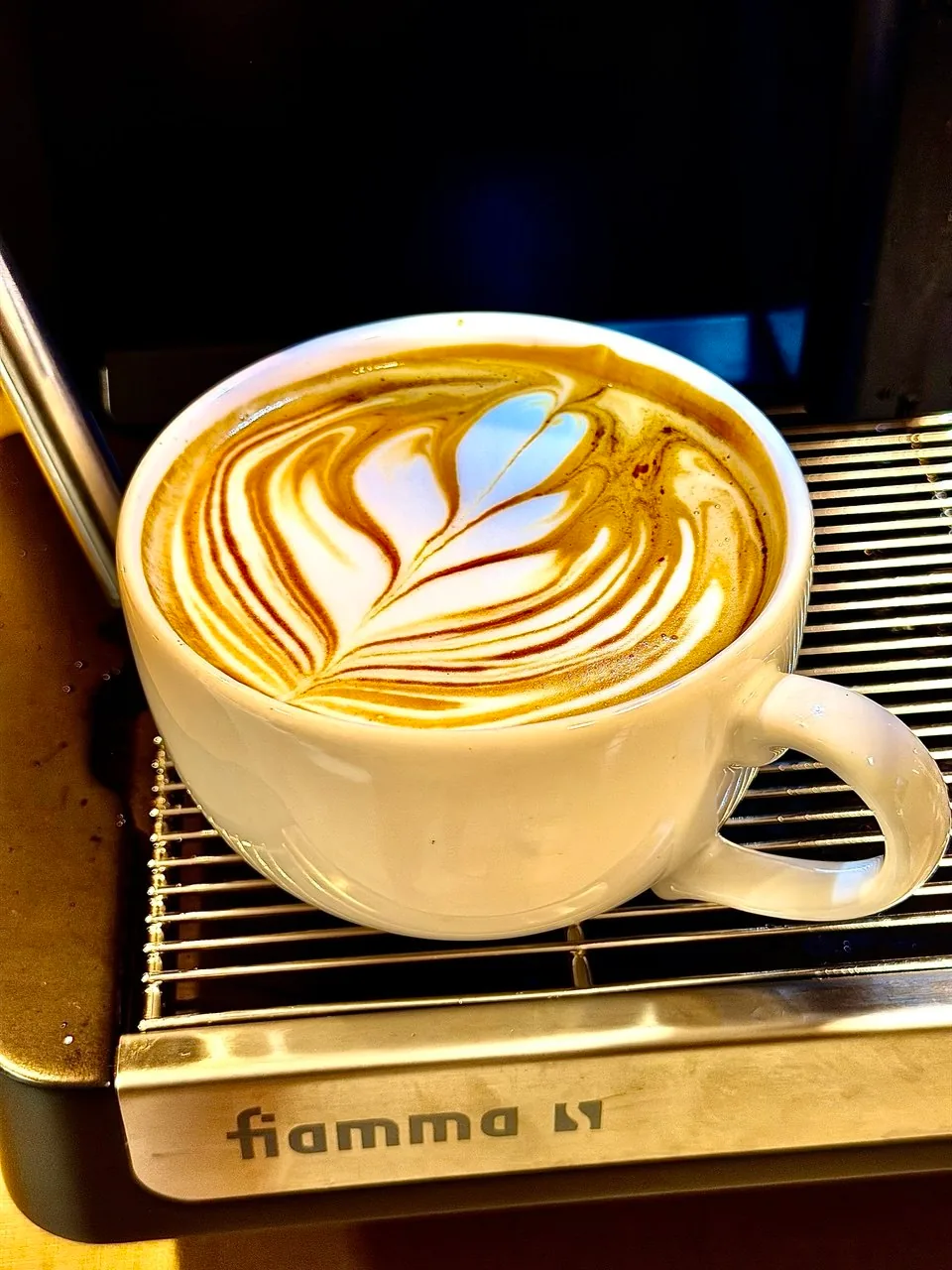 Xícara branca com latte art de folhas apoiada na bandeja de inox da máquina Fiamma.